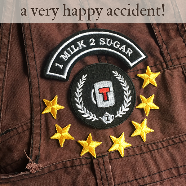 A close-up views of a pair of brown dungarees with black stitching. Placed on top of them are a round embroidered patch depicting a mug of tea inside a laurel wreath, a curved patch above it that says "1 milk 2 sugar", and there are seven small gold star patches underneath.